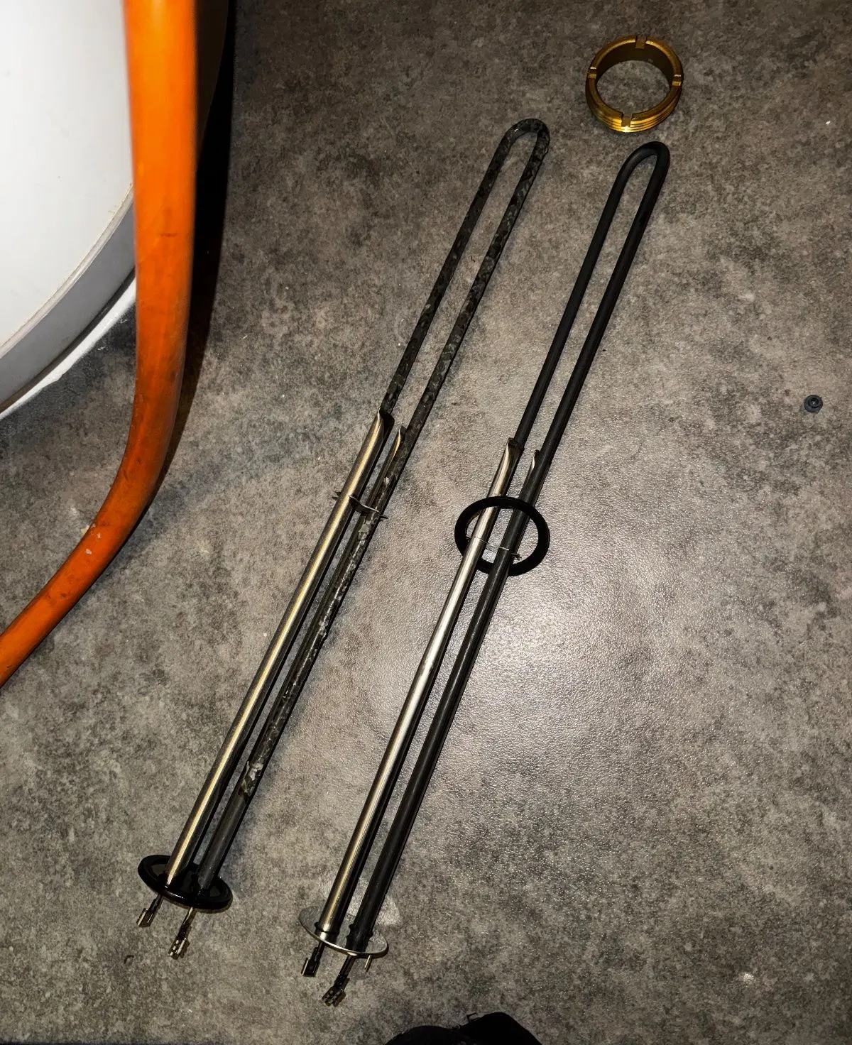 A removed immersion heater element with a dark crust of limescale, laid on the floor beside a clean replacement element